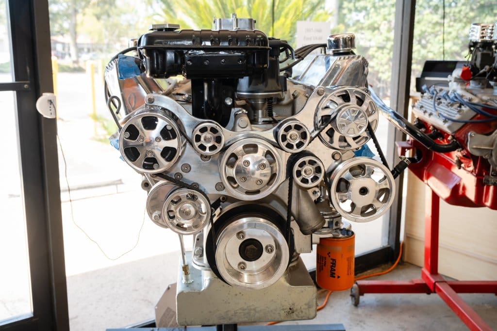 A metallic car engine displayed indoors, featuring various pulleys and belts arranged on its front.
